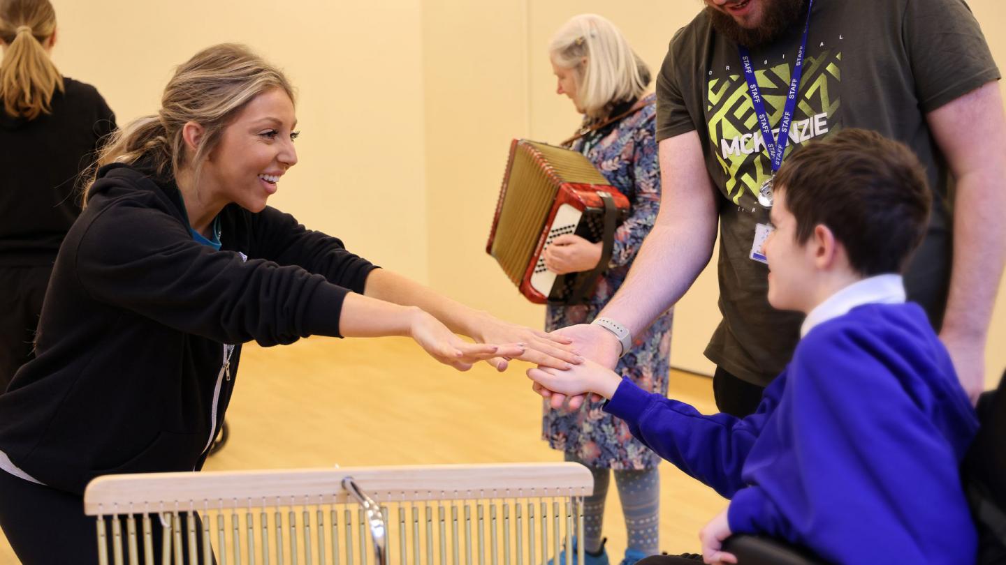A woman wearing a black top dances with a boy in a wheelchair wearing a blue school uniform, musicians can be seen playing in the background
