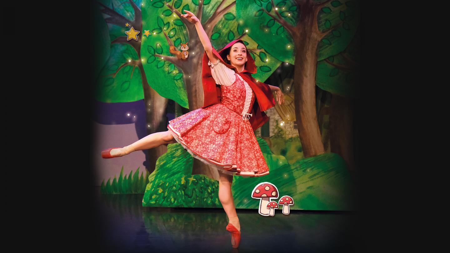 Dancer in red with a red hood and red pointe shoes stands arabesque smiling as she walks through a forest