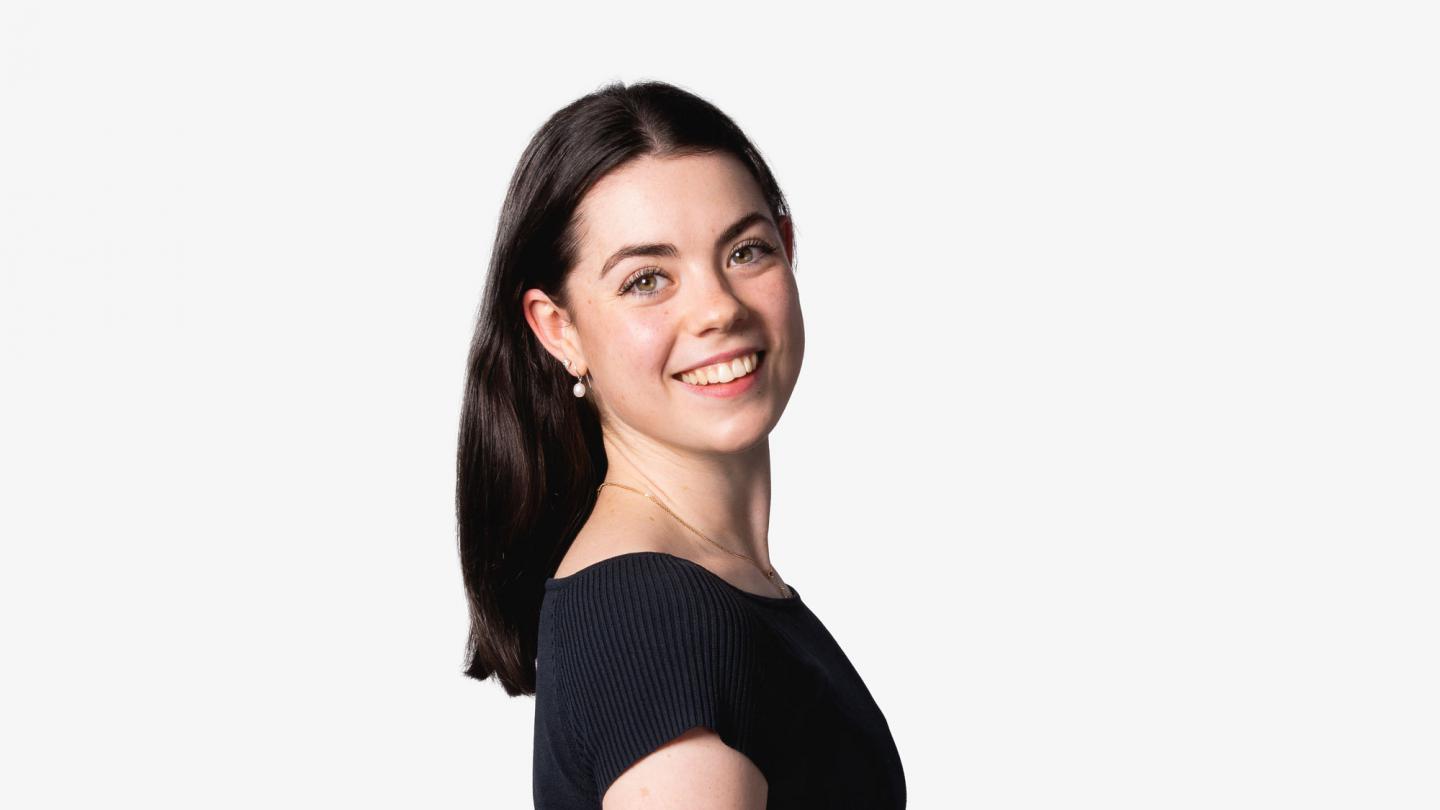 A dancer stood on a white background with medium length brown hair smiling to camera