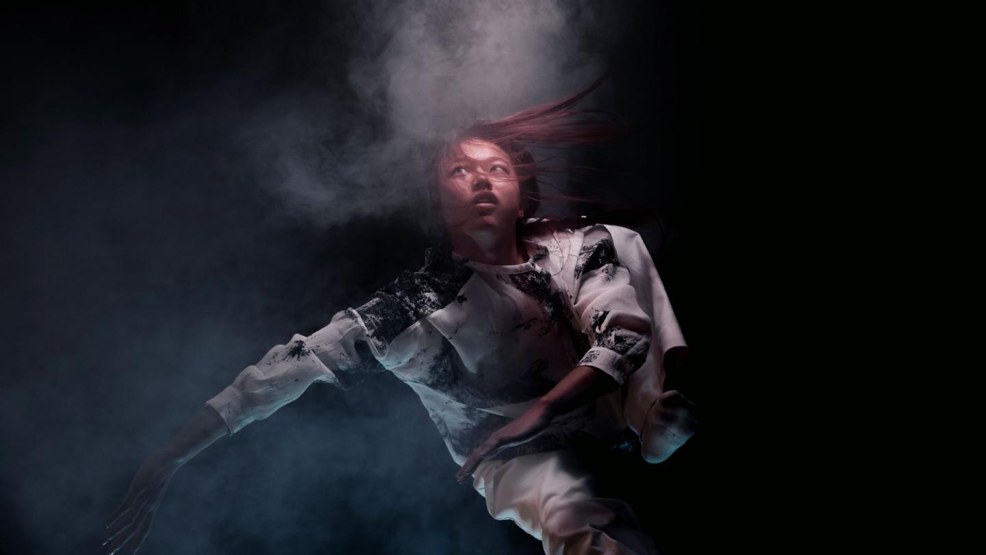 On a black background, a dancer is captured in mid-motion, with their body suspended in the air and surrounded by a swirl of smoke. 