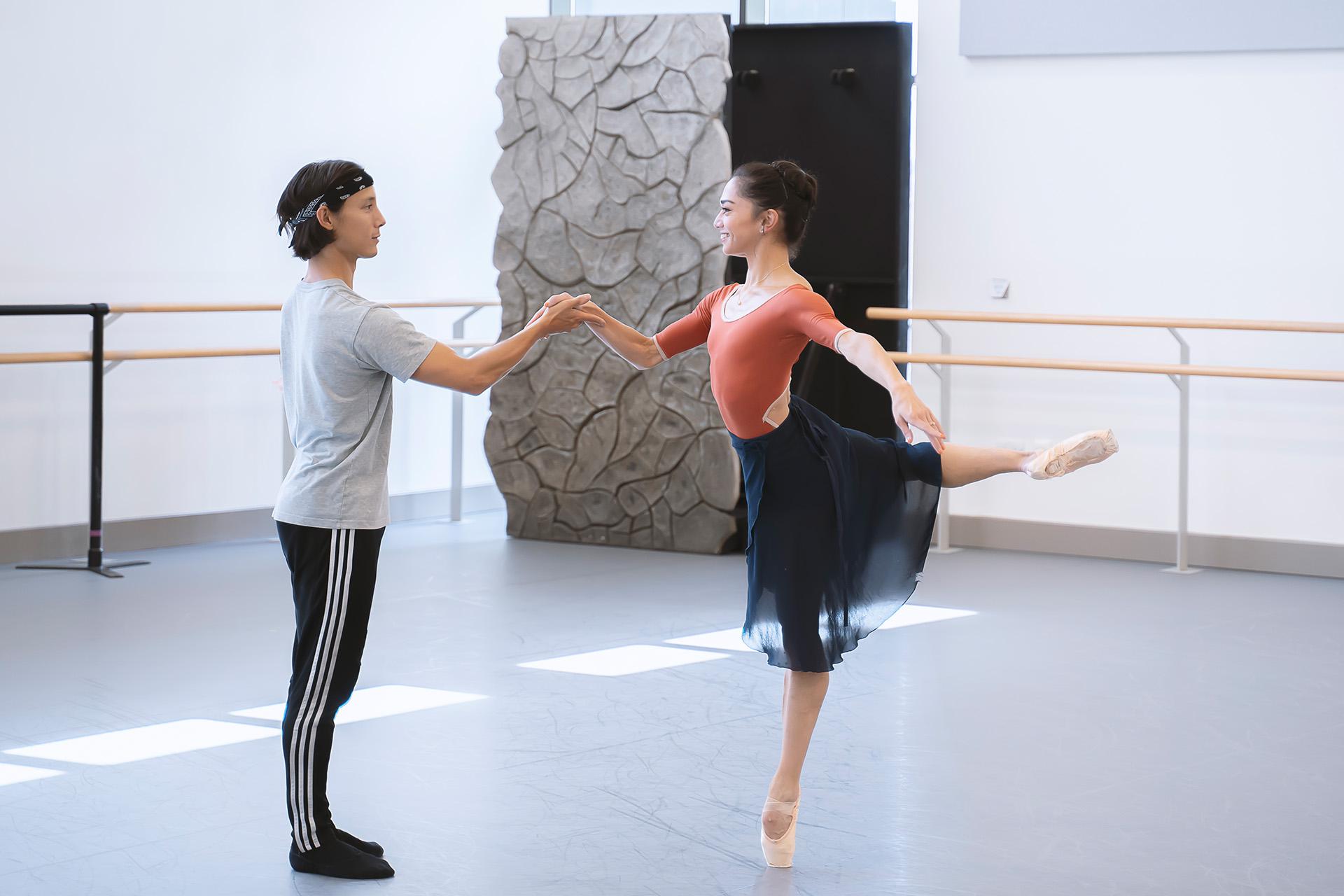 A dancer holds another hand to keep them steady as they balance on a pointed foot, their other leg bent behind them