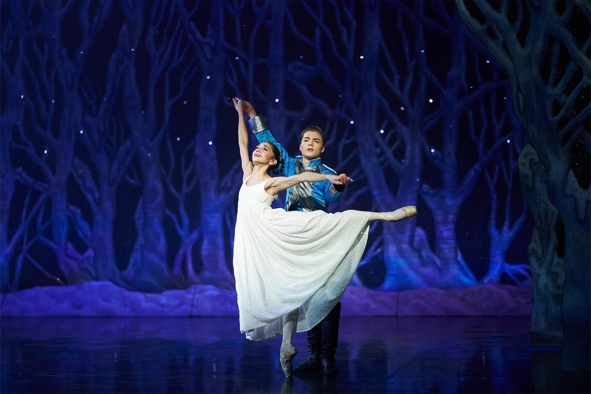 Dancer wearing a white dress stands in a snowy scene supported by a soldier in full dress uniform