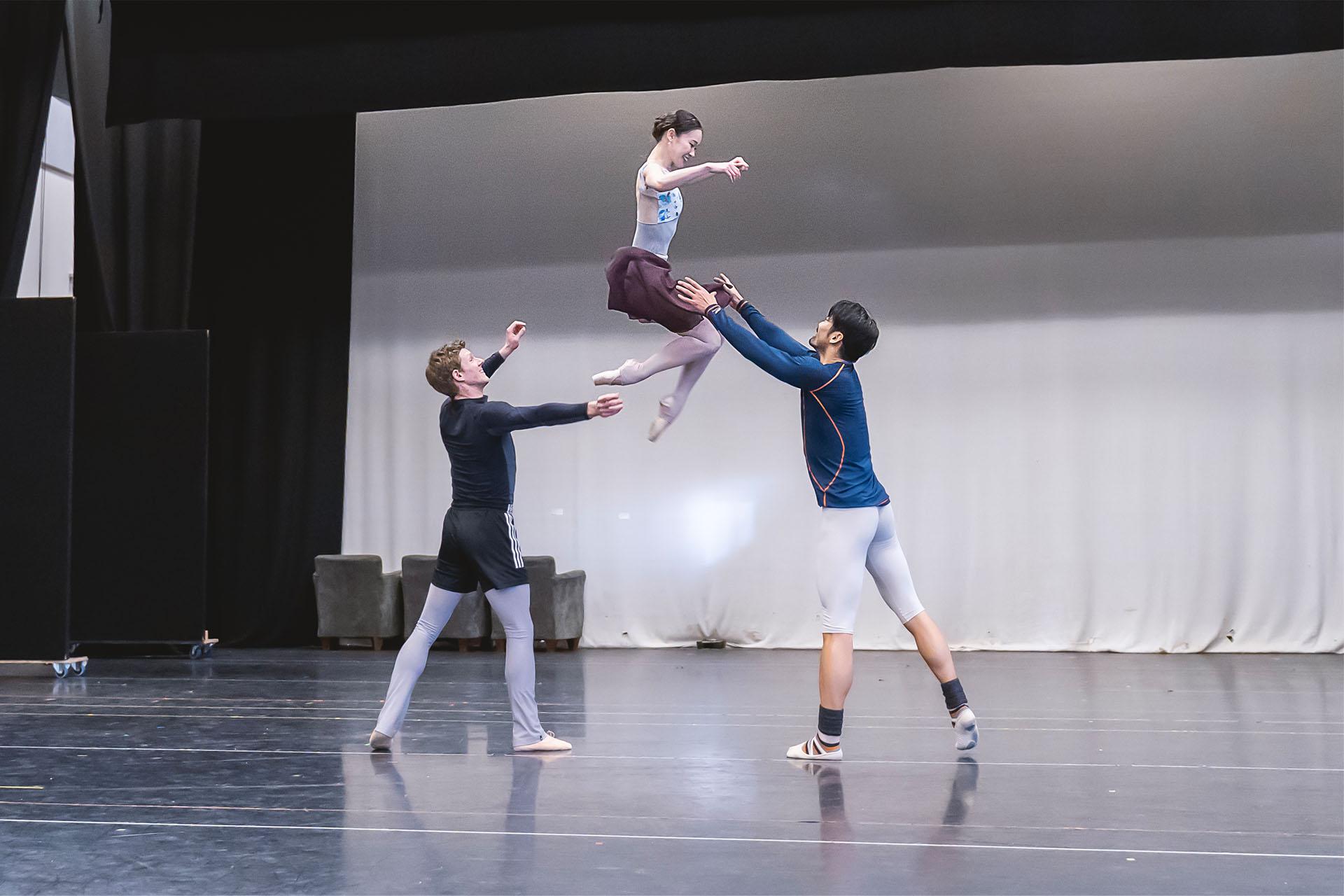 Three dancers rehearsing, the woman in midair as the two male dancers pass her from one to the other