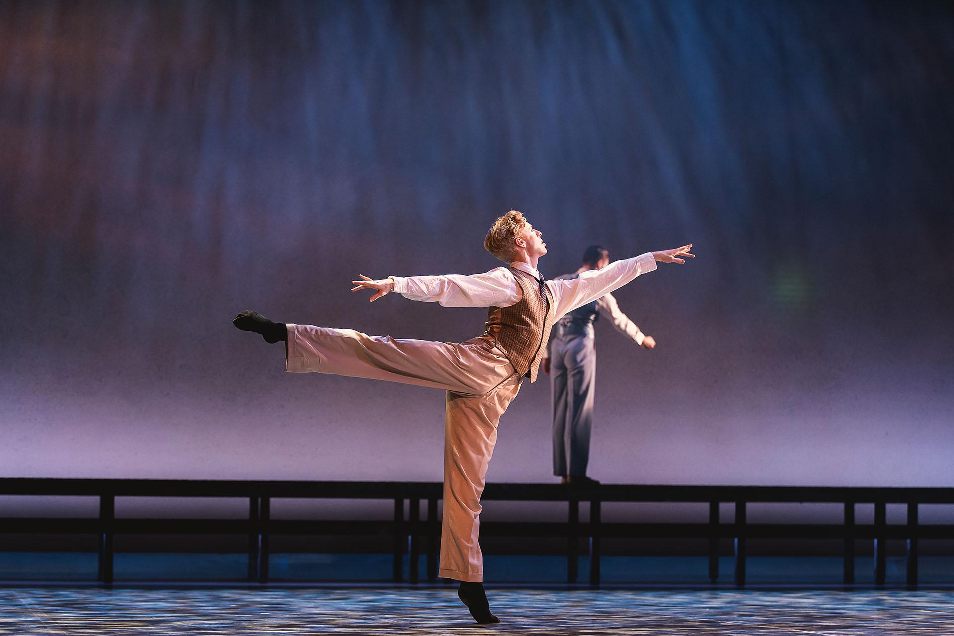 Male dancer in beige trousers and waistcoat and a white shirt stands arabesque