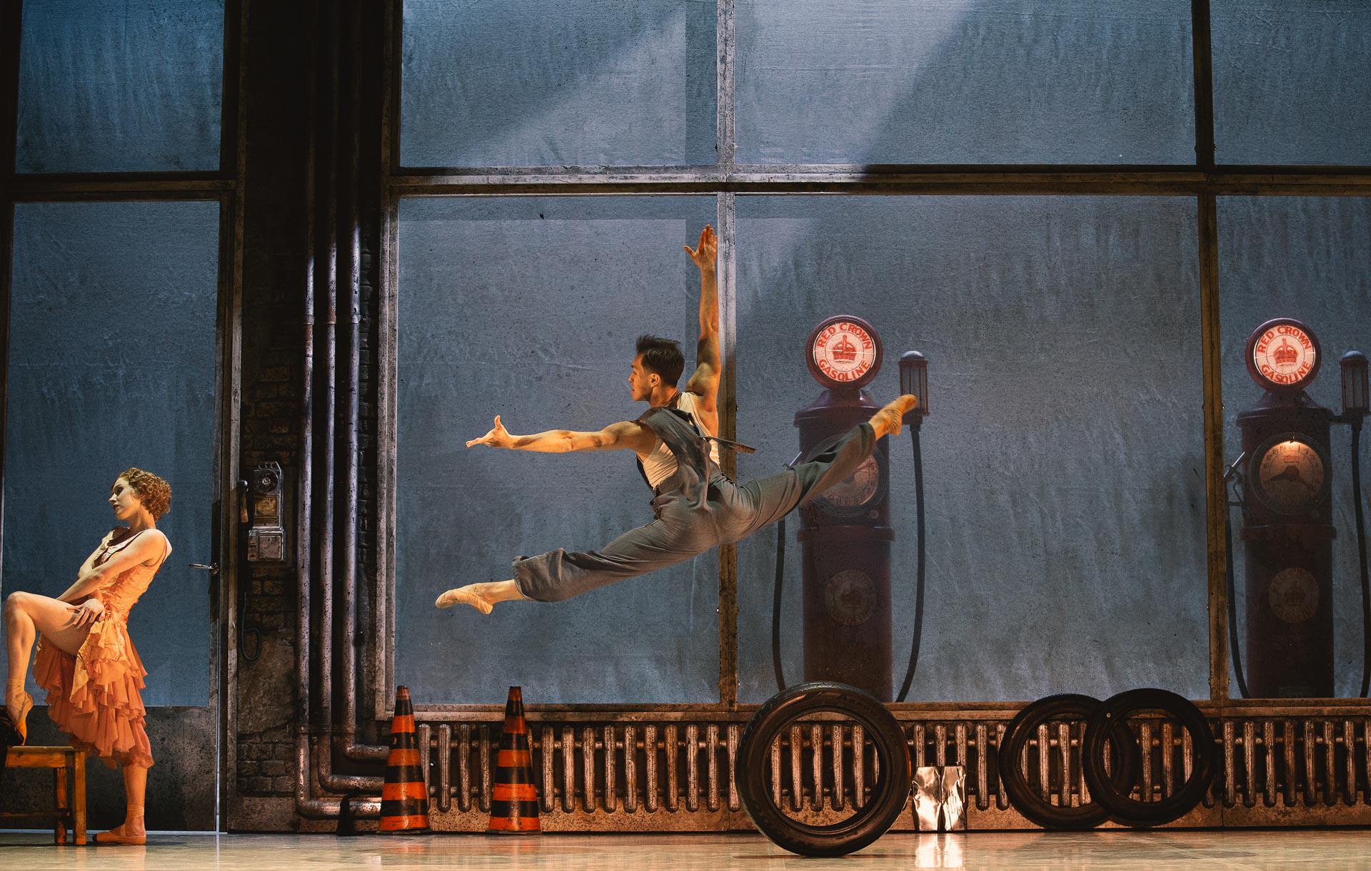 A male dancer jumping in splits on stage set like a car garage