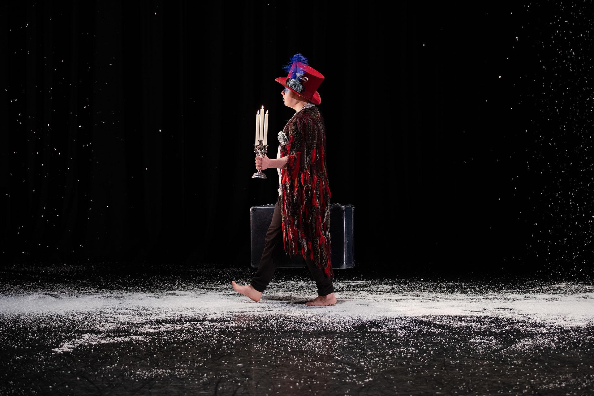 PErformer in a red top hat moving across a dark stage covered in snow