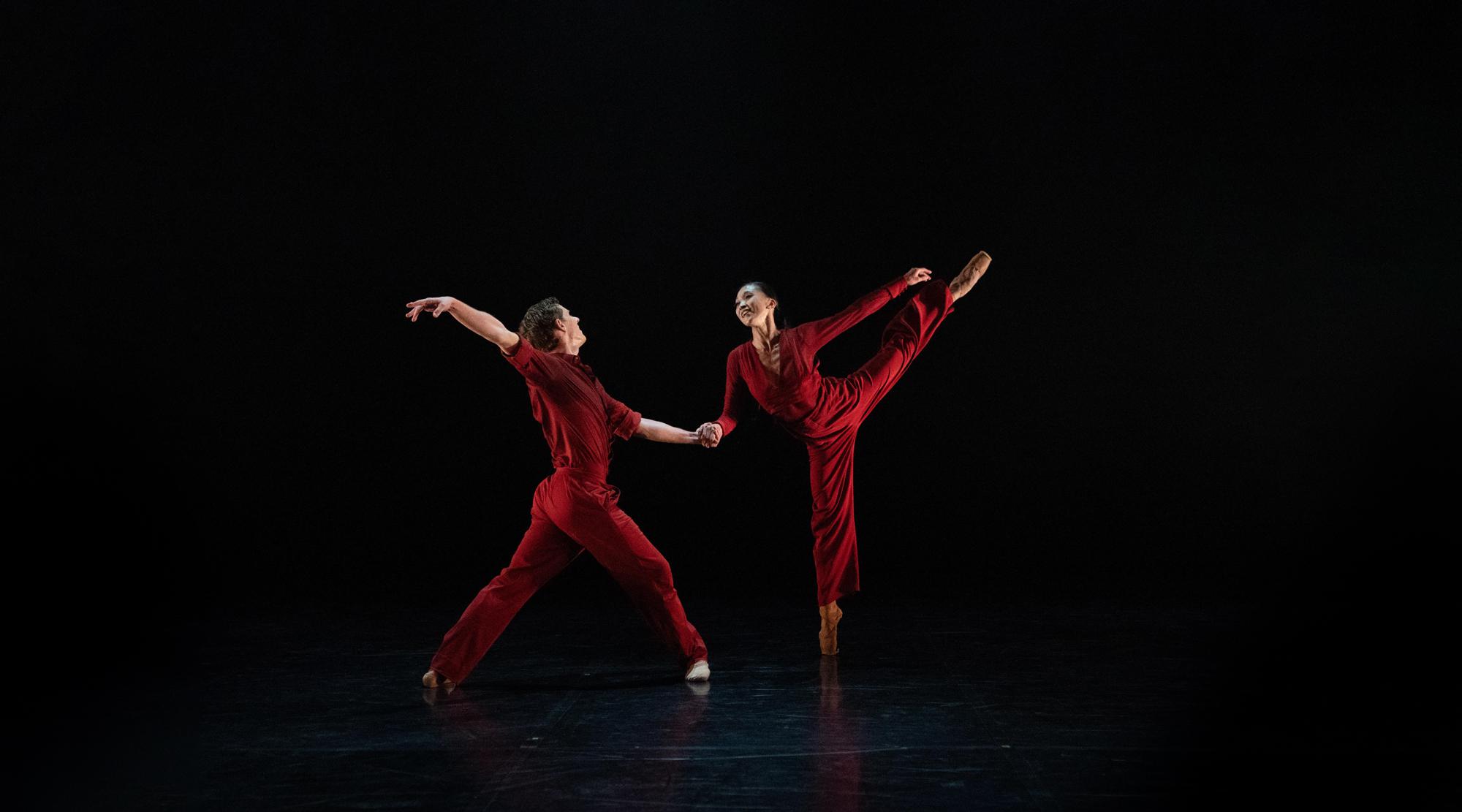 Male and female dancer in red jump suits standing on point with one leg raised high.