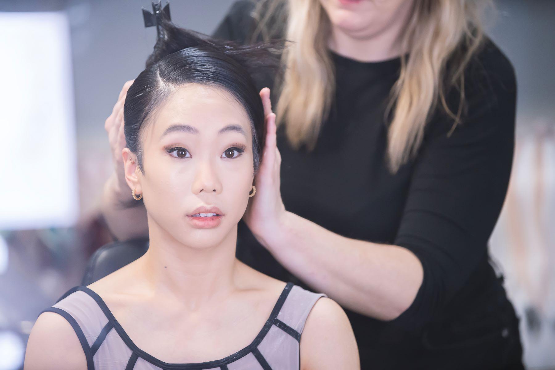 A female dancer looks past the camera as a stylist adjusts her hair.