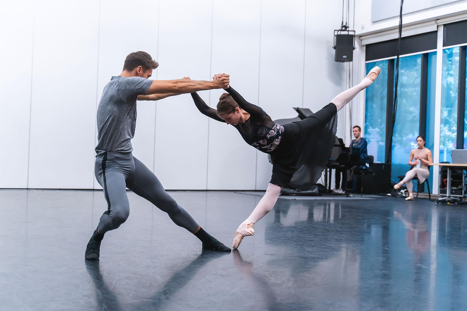 Female dancer en pointe in extreme arabesque, her extended leg behind her raised and her body leaning forward as a male dancer supports her by her arms which are raised above her head.