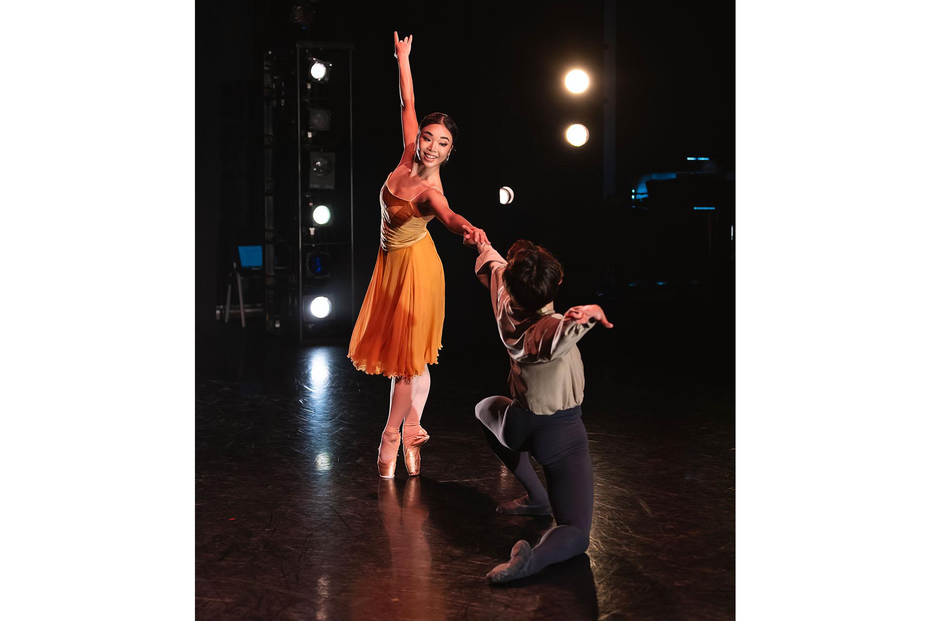 Female dancer wearing a vibrant orange dress stands on her toes and reaches across to a male dancer in grey tights and shirt who is kneeling on the stage and holding her extended hand