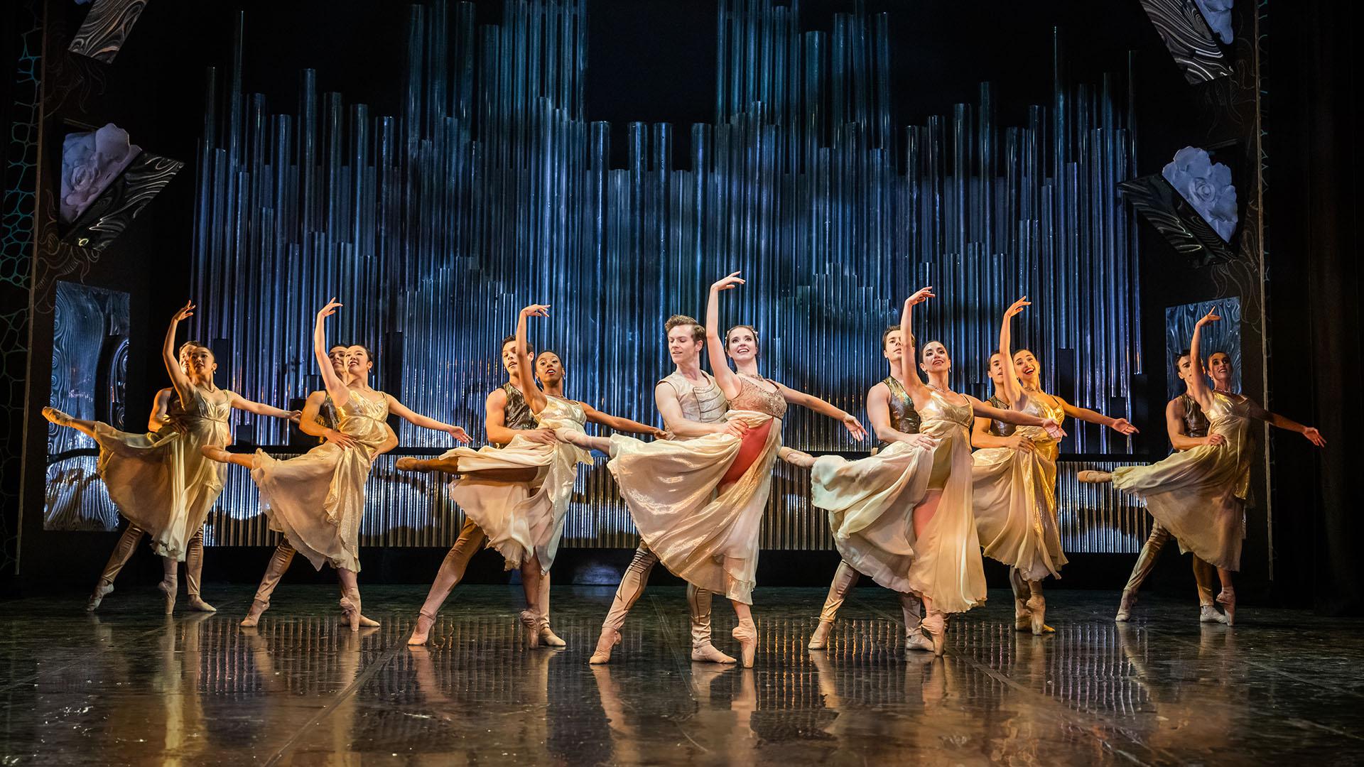 Dancers, dressed in pale gold costume, paired with the male holding the female by the waist for stability as she in en pointe ion one foot and in arabesque.