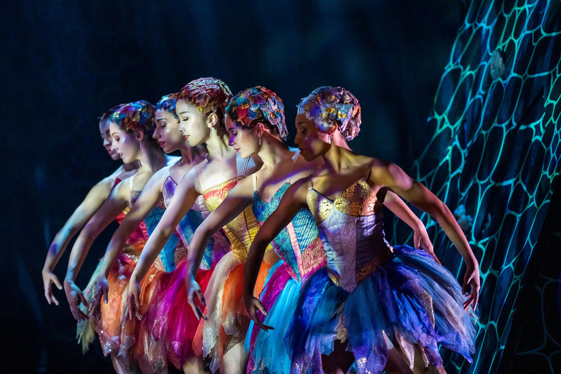 Six female dancers, each wearing brightly colourful dresses and hats stand in a row looking down