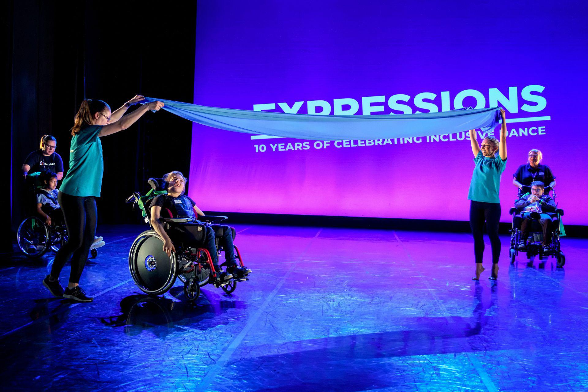 A group of children perform a wheelchair dance routine with their support workers on purple lit stage