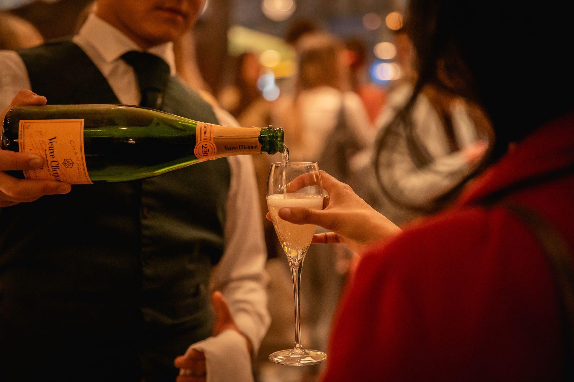A man pours champagne into a woman's glass, a gathering of people can be seen behind them.