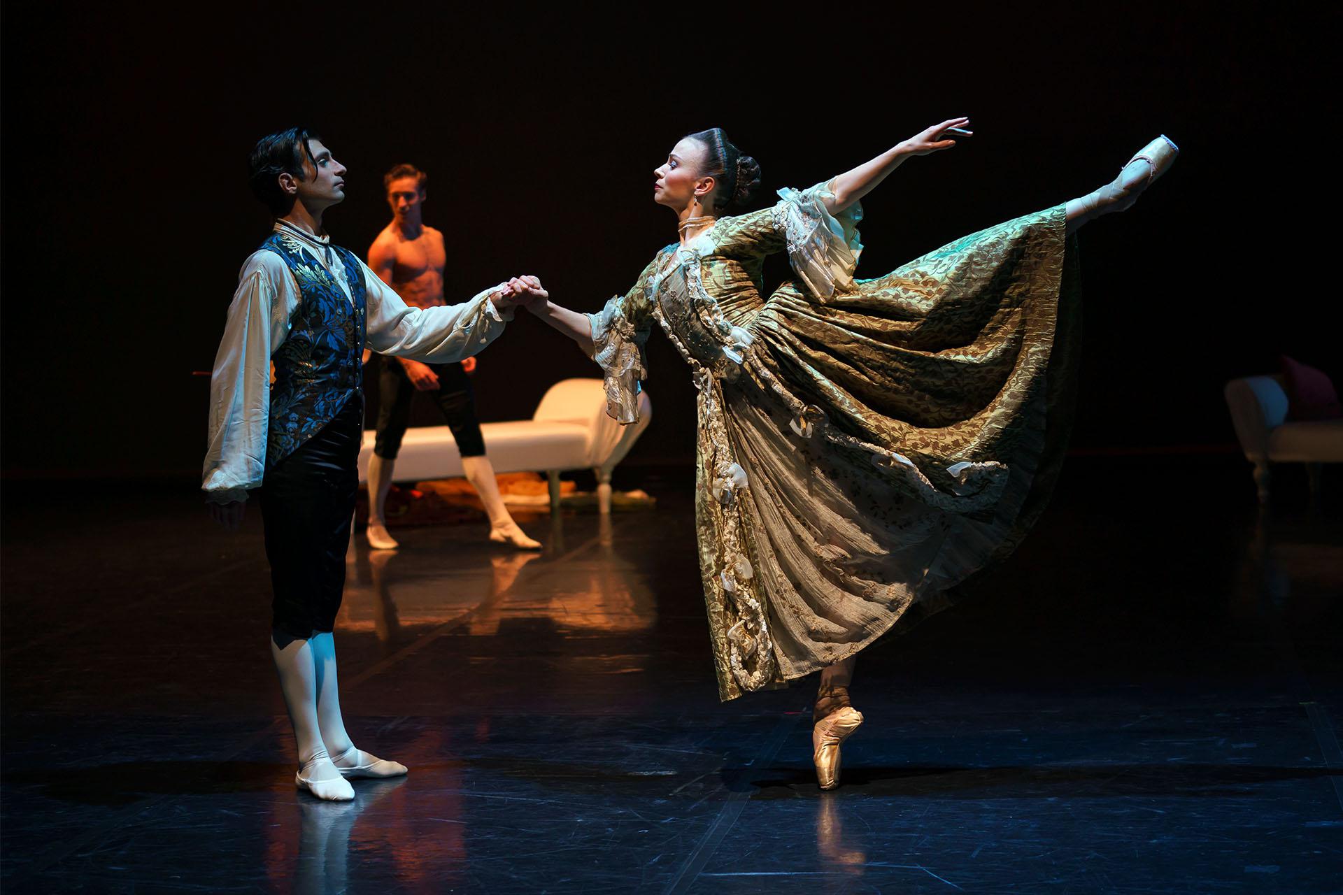 Dancer stands on one foot en pointe, a leg raised behind her arabesque, she is wearing an intricate 18th century gold costume