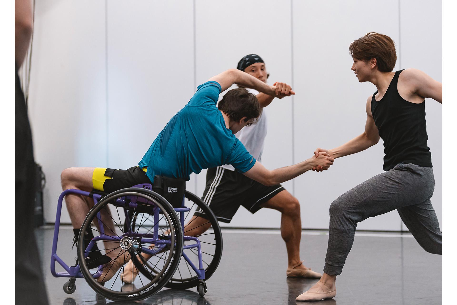Wheelchair dancer leans back and grabs the hands of two other dancers, one beside him the other behind