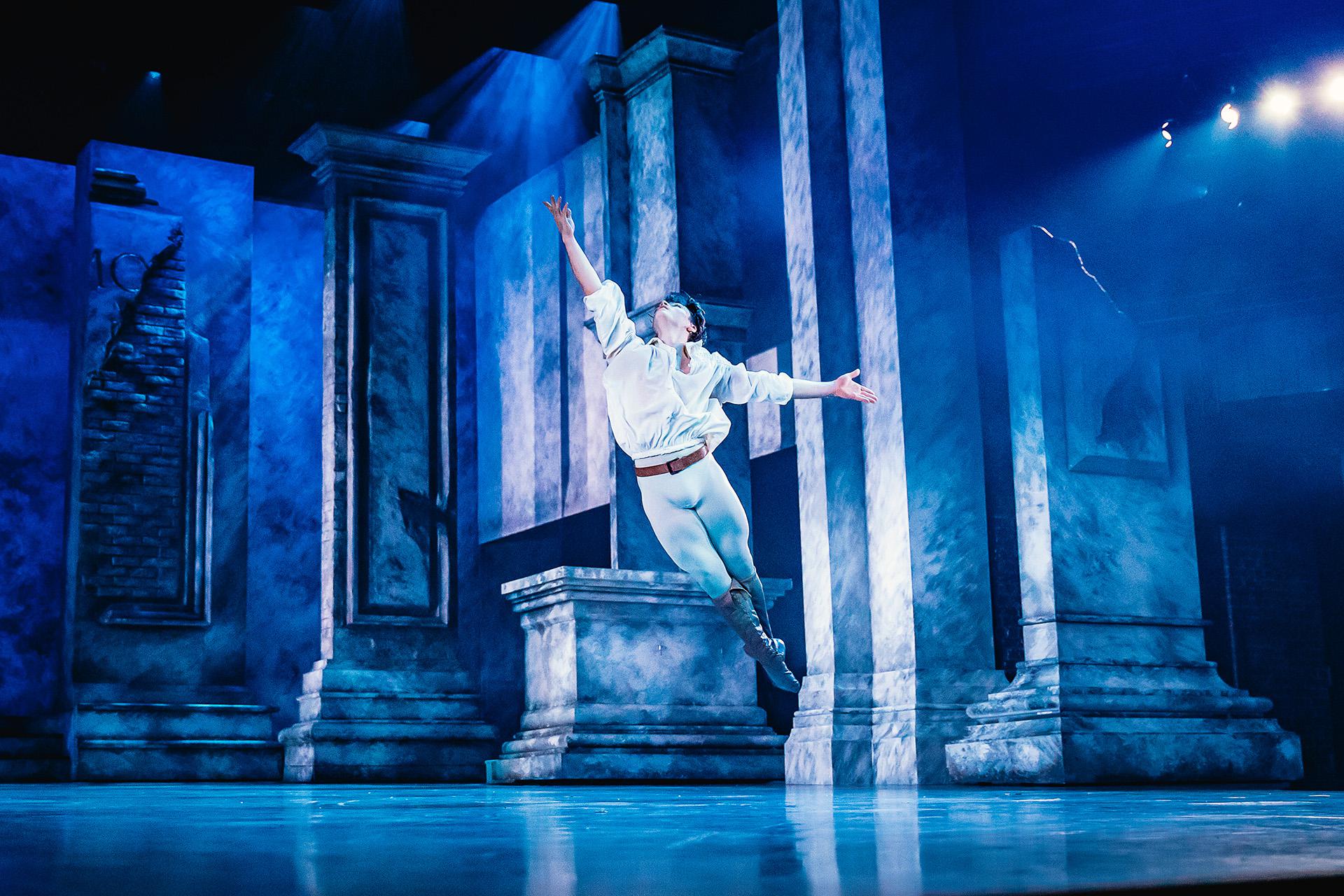 Dancer leaping across the stage, his legs together, one arm raise triumphantly above him, bathed in blue light