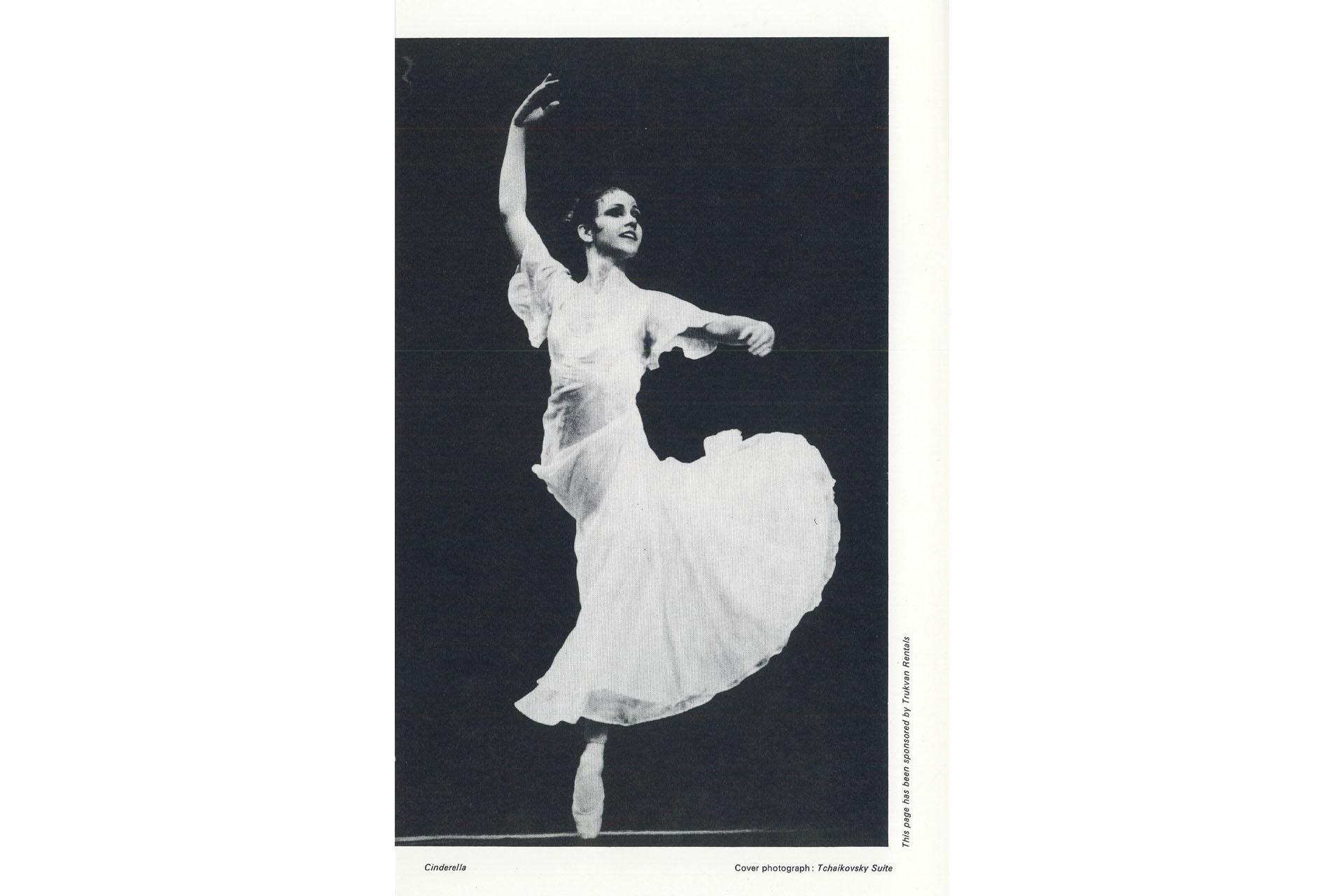 Black and white photo of a dancer in a long white skirt stood on one leg