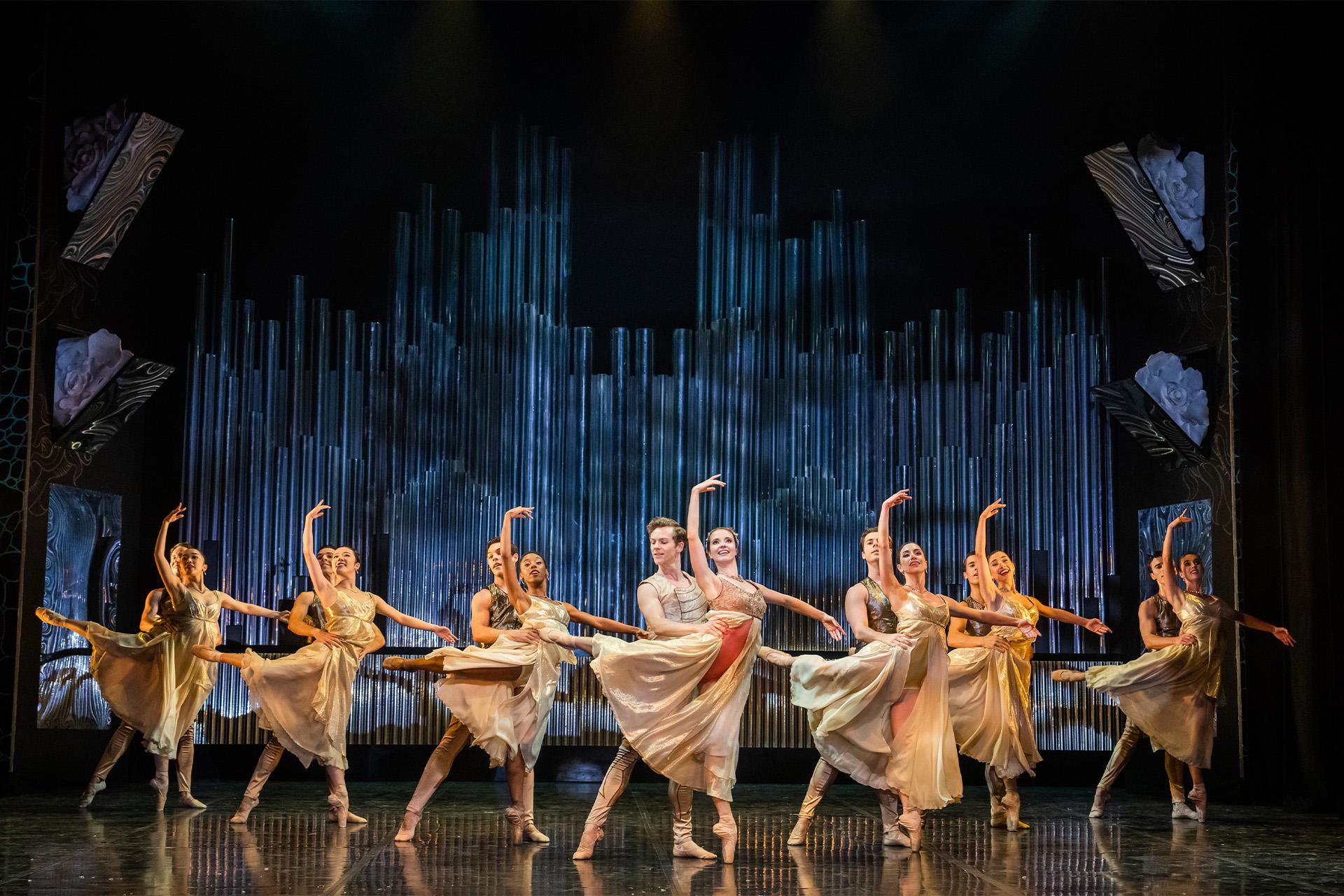 Fourteen dancers all stood together in couples. One dancer in each couple has their arm raised and standing on one foot. Lots of gold and blue colours