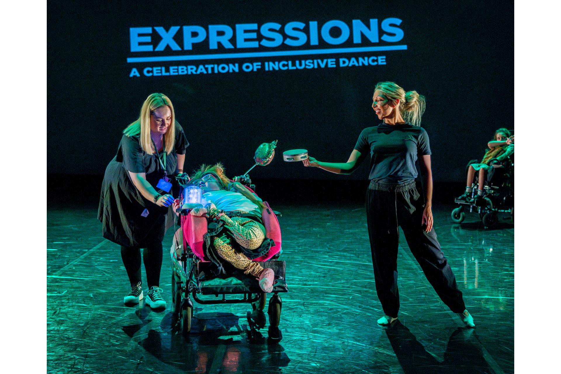 A dancer in black hands a tambourine to another dancer in a wheelchair.