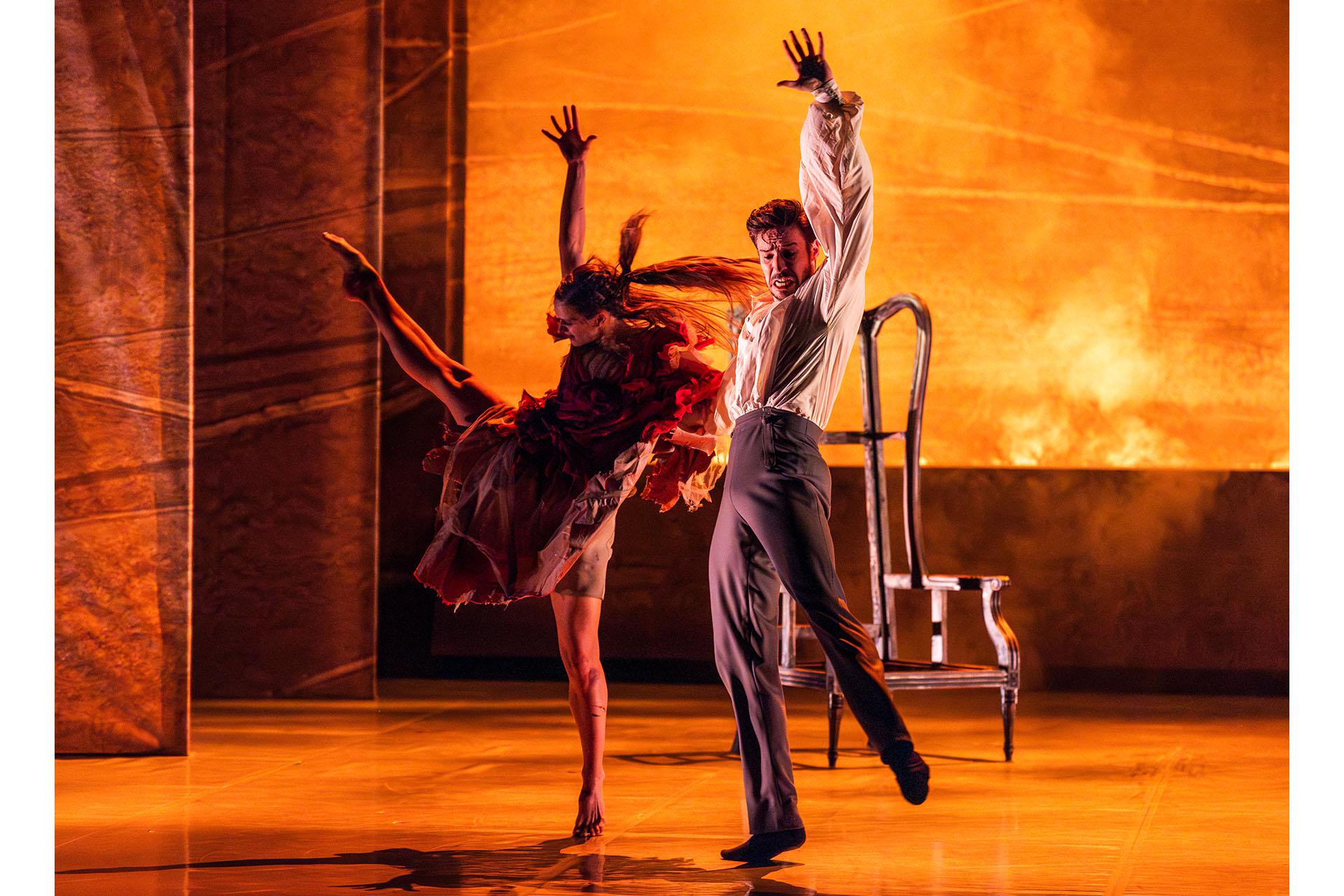 While an inferno rages behind them, a dancer in a red dress performs wildly while a male dancer is behind her, angry and trying to control her