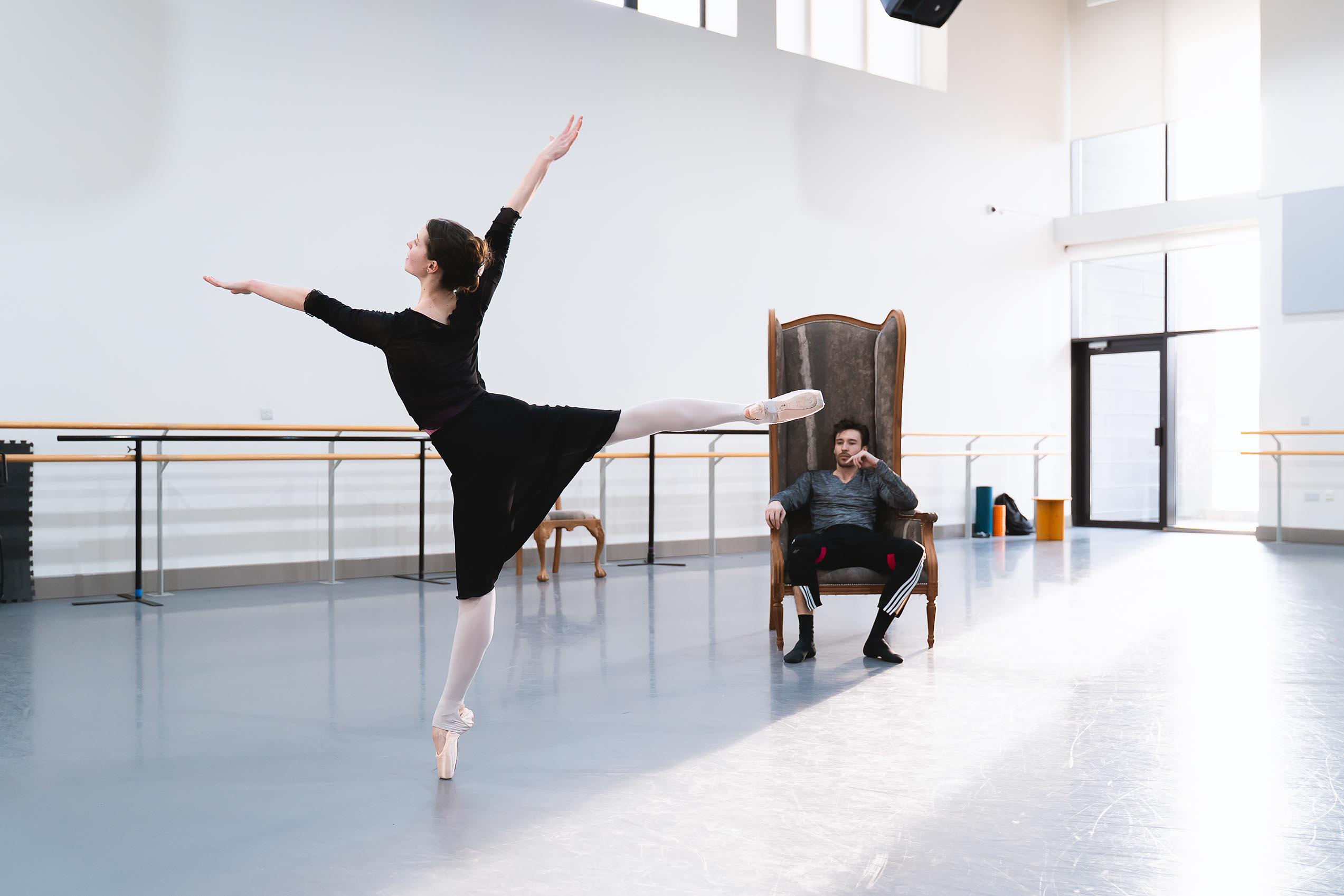 A dancer stood on pointe with legs and arms outstretched with another dancer watching on in a large armchair