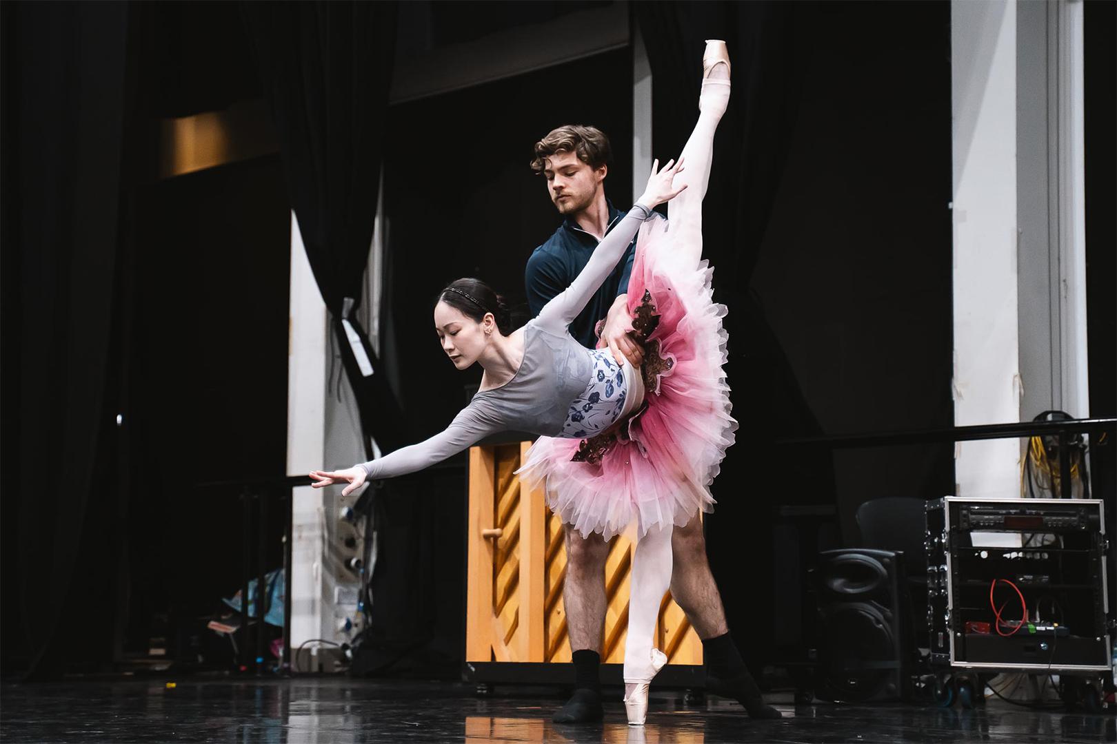Two dancers in shot, one behind the other. The dancer in front leans forward on her toes wearing a pink tutu and reaching down.