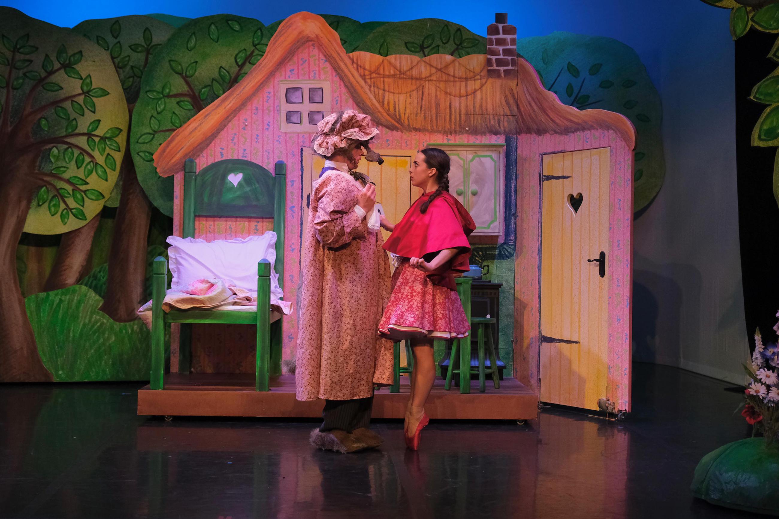 Two dancers stood on stage facing each other, one is dressed in a wolf costume and the other is wearing a red dress and cloak