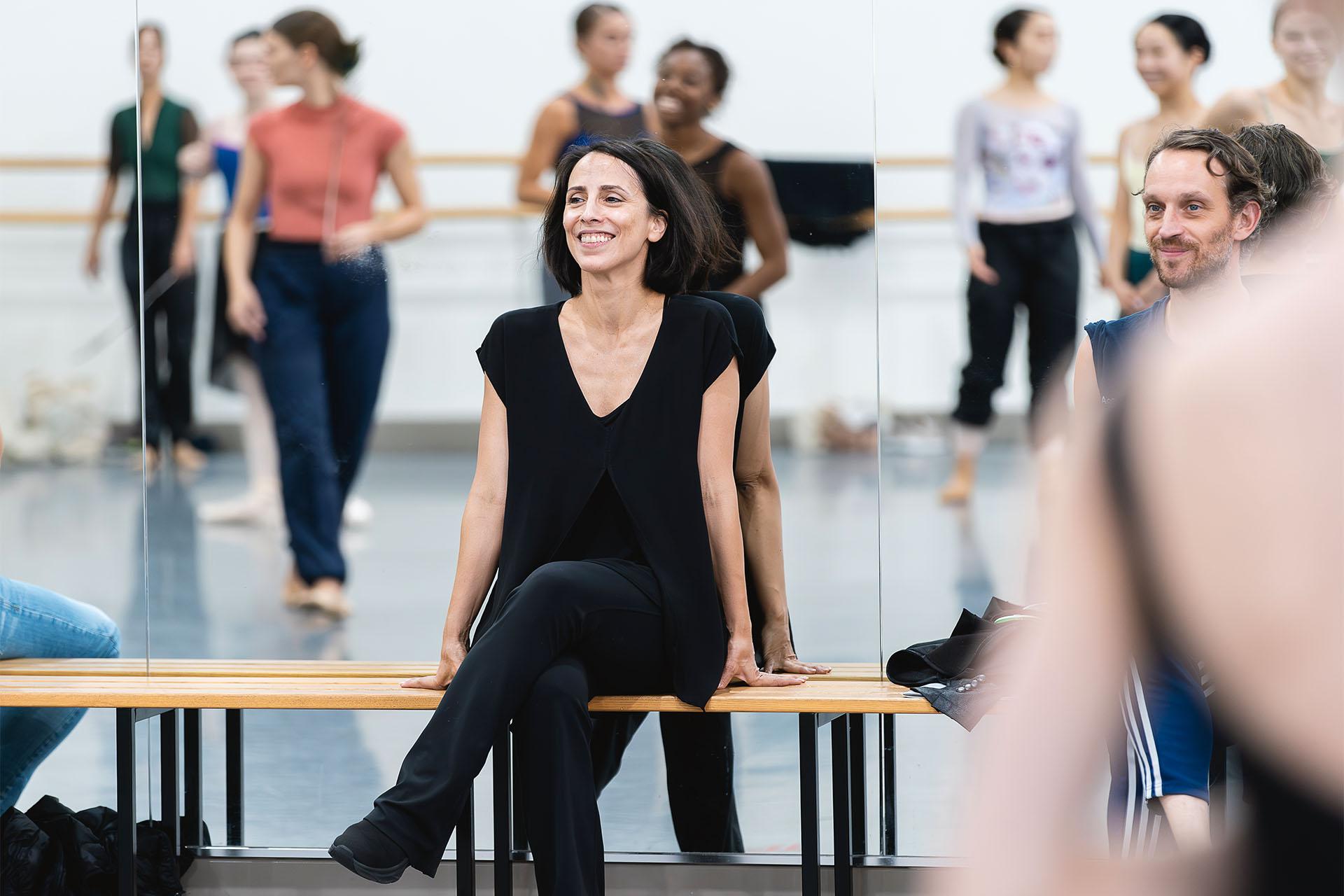 Woman in black sits on a wooden bench in front a full length mirrors smiling. In the reflection are dancers also smiling.