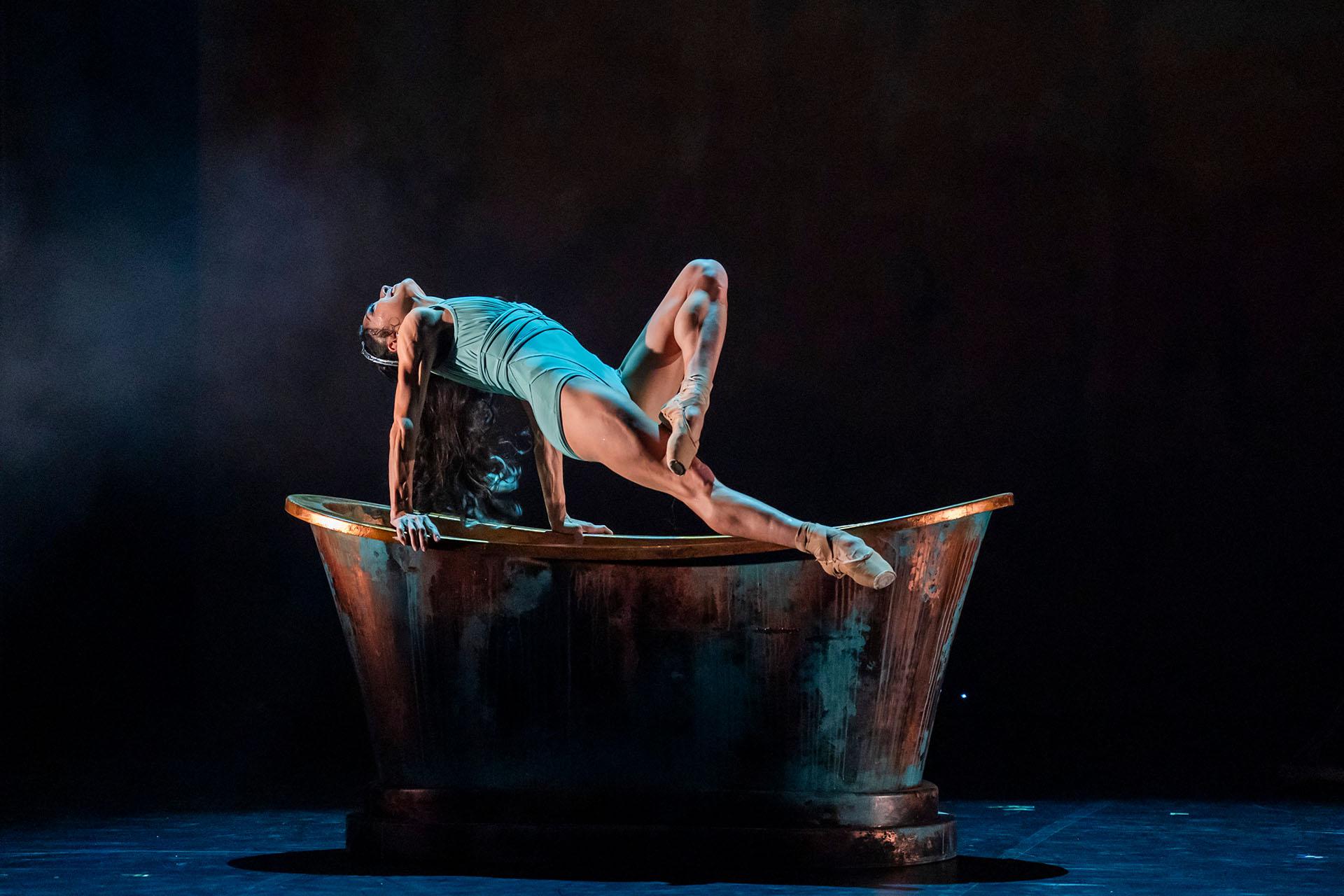 Ygraine wearing a turquoise shift lifts herself above a copper bath, her back arched, her head thrown back