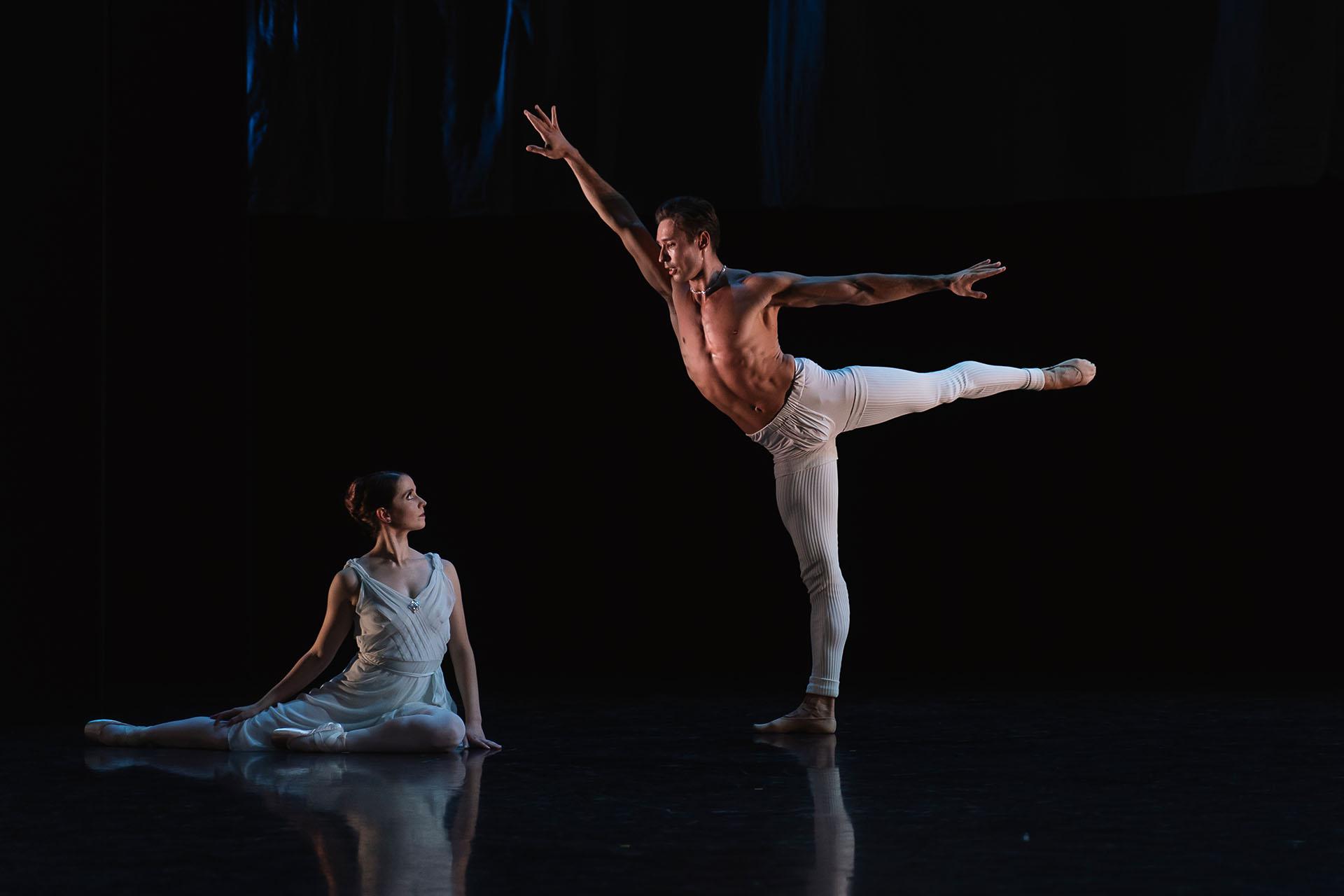 A woman sits on the floor looking a a man standing on one leg, his other outstretched behind him, his arms out for balance