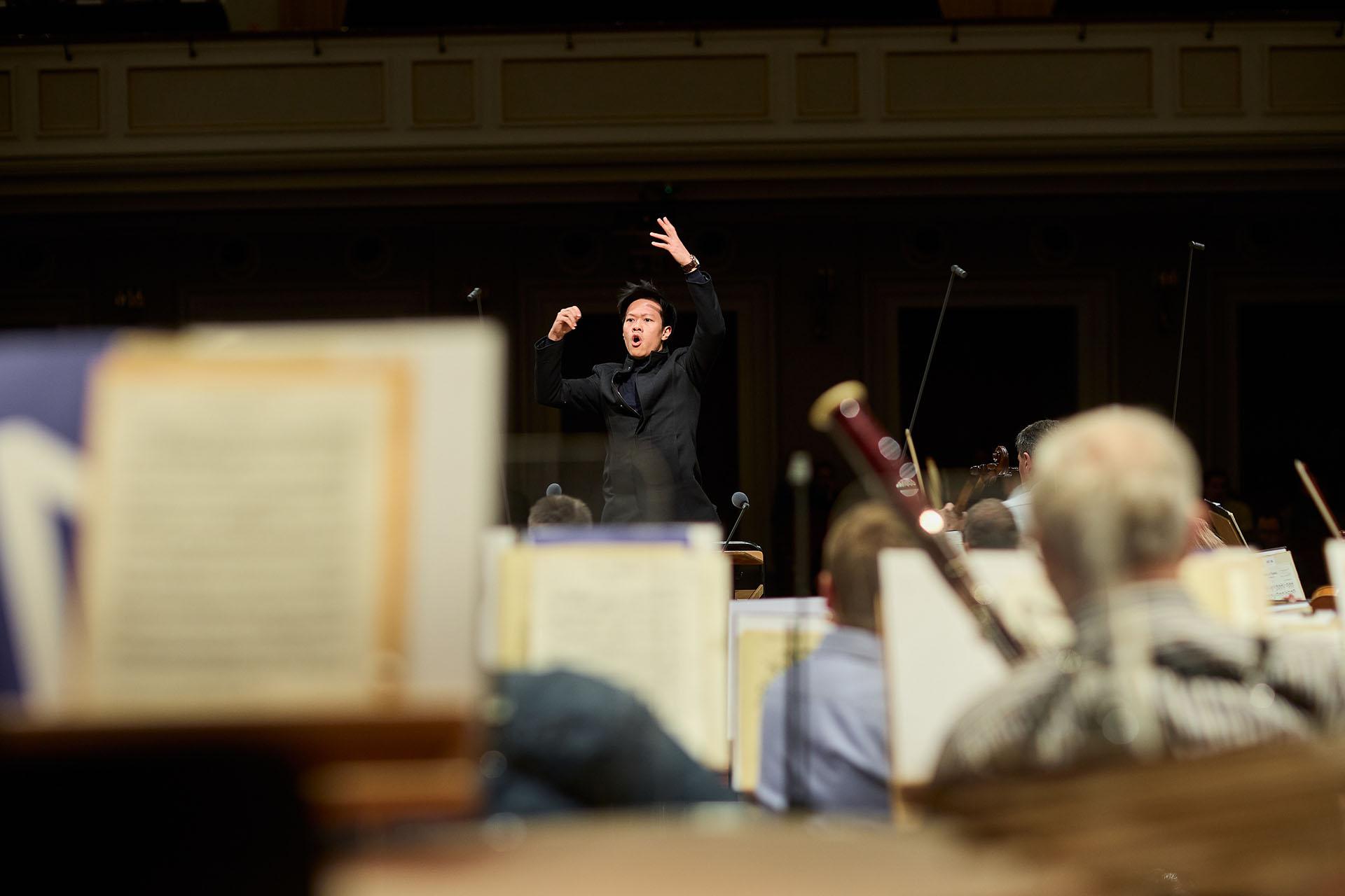 Conductor with a raised hands wearing black, viewed over musicians