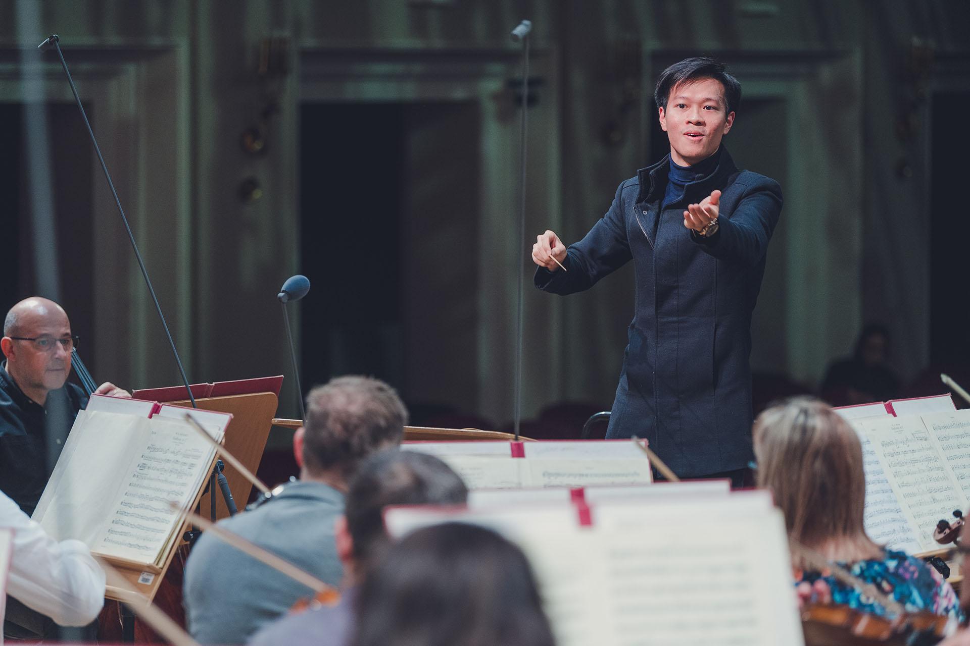 Conductor wearing black, left hand raised to the musicians, right hand holding the baton