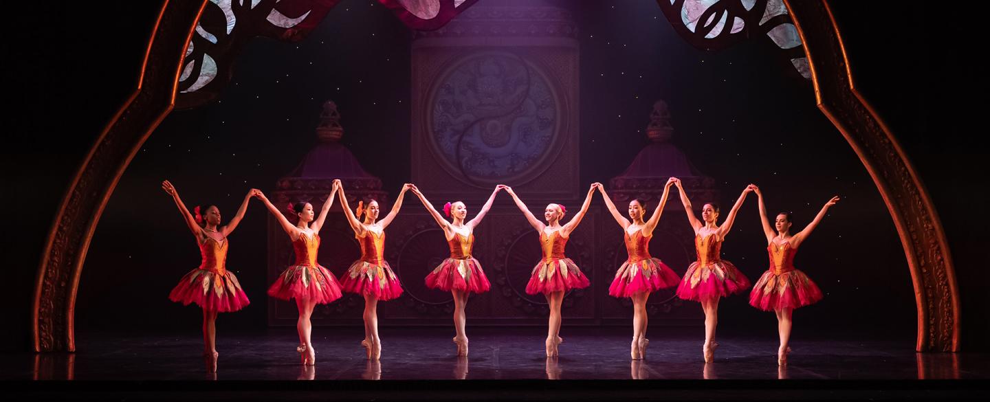 Dancers all dressed in pink and orange bodices and skirts stand in a line on their toes and arms high in the air