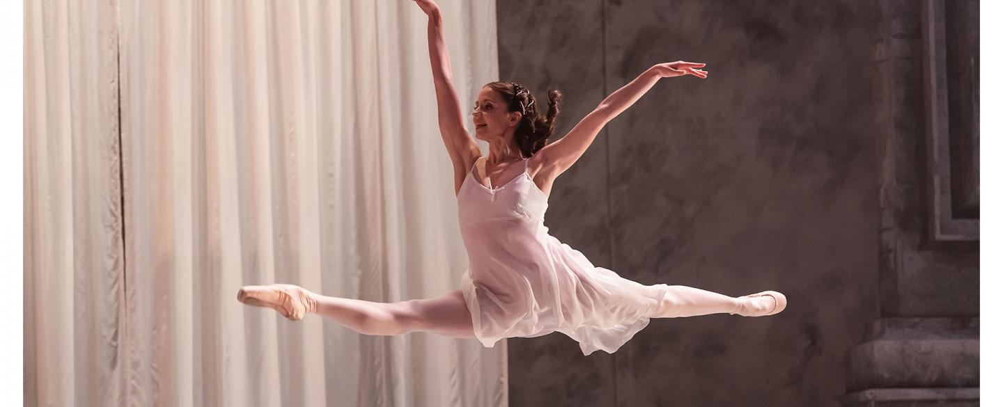 Dancer wearing white flies across the stage, leaping in a grande jete