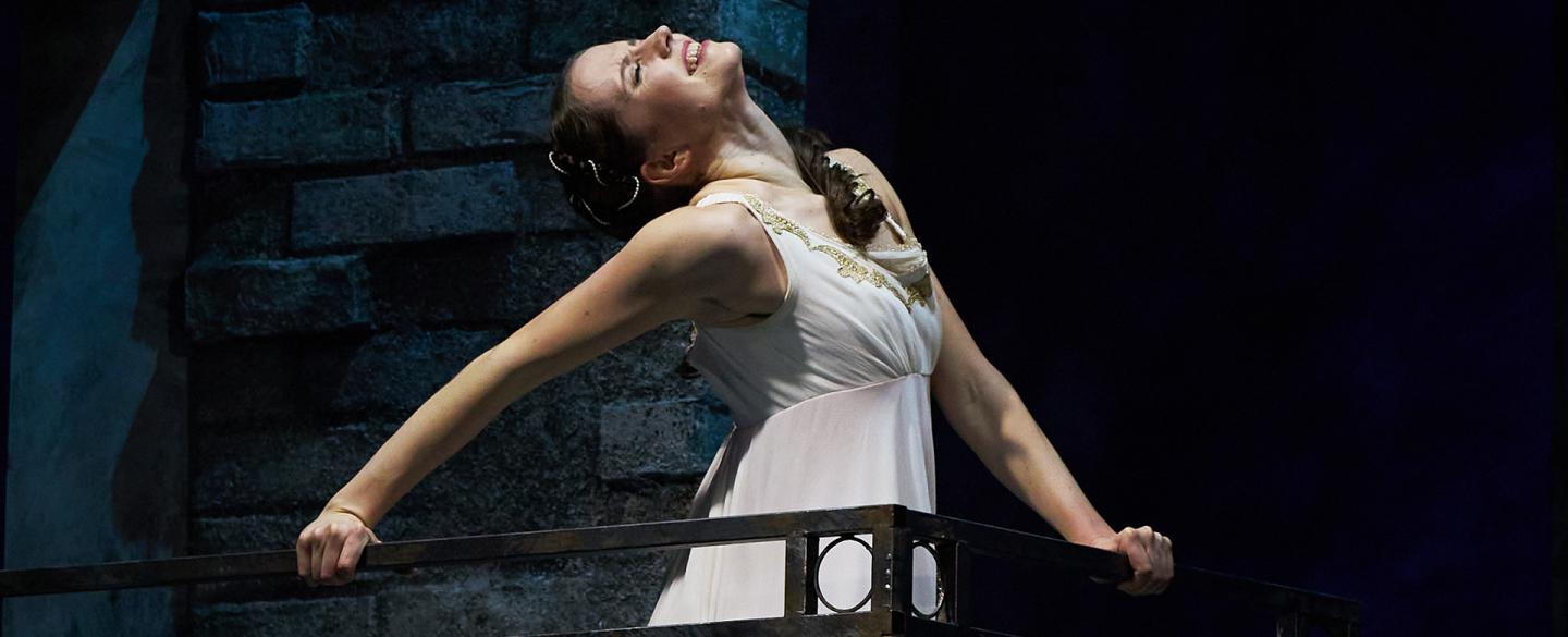 Juliet in a white dress standing on her balcony, her head back, eyes closed and smiling with ecstasy
