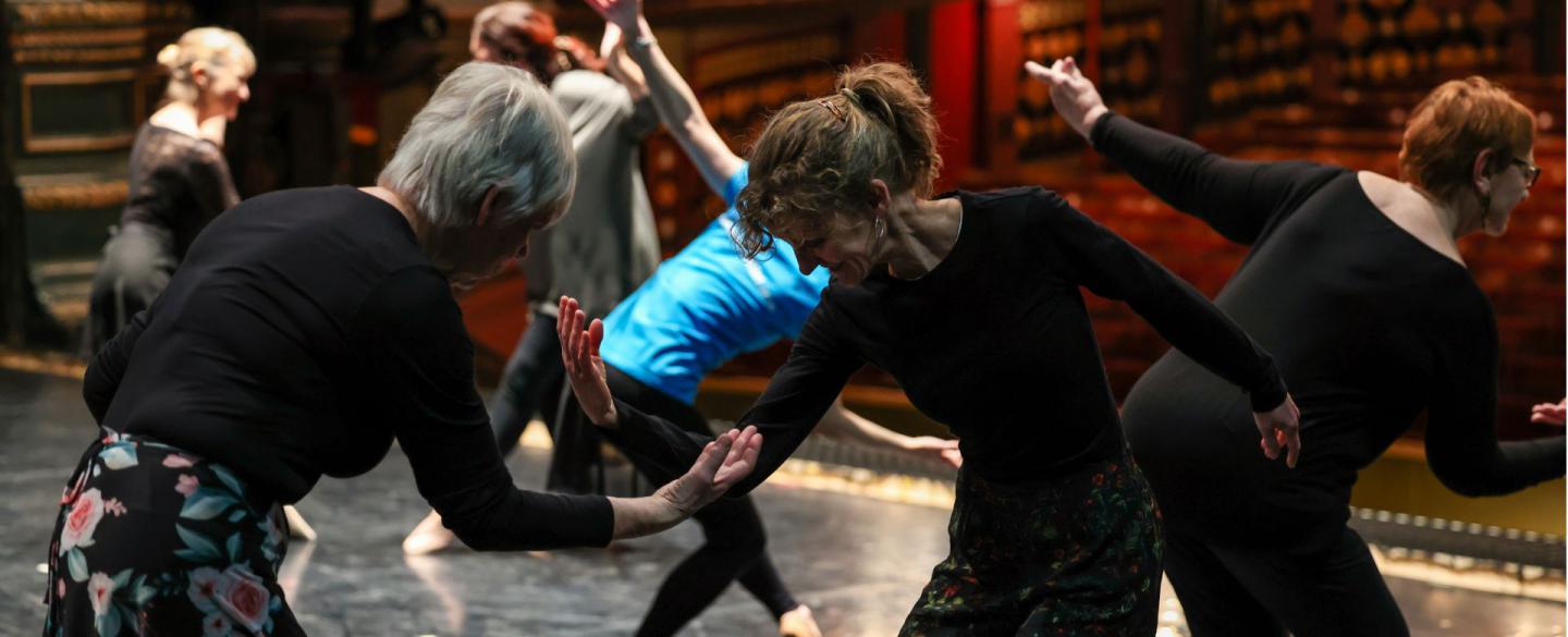 Older dancers rehearse ballet moves on a stage. Two stand in front of one another, bend forward and begin to lift their arm towards the other