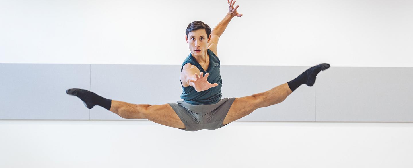 A dark haired dancer leaps, legs spread mid-jump, one hand outstretched to the camara