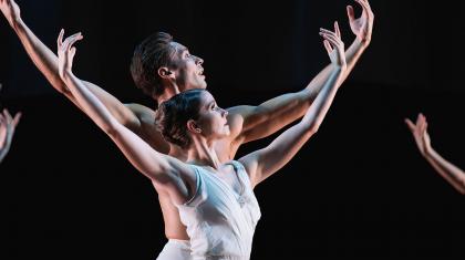 Dancers in white standing one behind the other, half look to the sky with their arms raise like Atlas