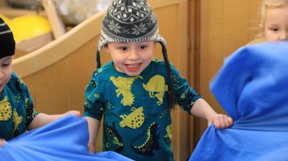 A young child smiling wearing a wooly hat and a green jumper with yellow and black dinosaurs