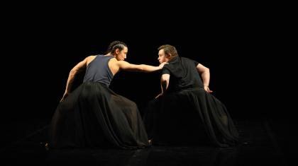 Two dancers stand hands on their bent knees. They look at each other as one grabs the other's shoulder.