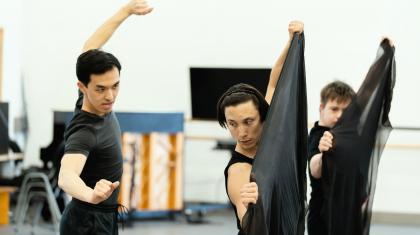 Two dancers hold pieces of black fabric in front of them, following the instructions of a third dancer.