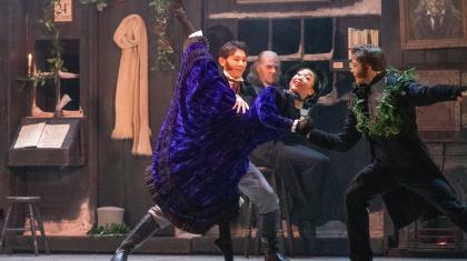 Scrooge's Nephew hold his wife by the waist as she stands on one foot en pointe and raises the other into the air