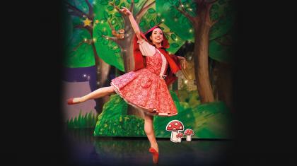 Dancer in red with a red hood and red pointe shoes stands arabesque smiling as she walks through a forest