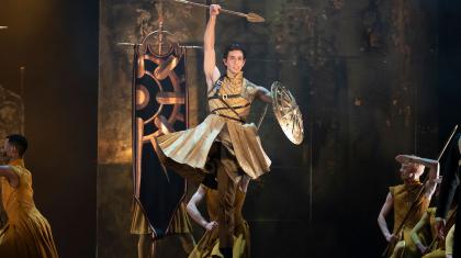 Jumping, arm raised with a pike, the other arm holding a round shield, his clothing golden, Uther battles on